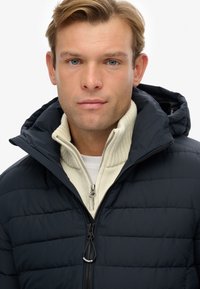 Chaqueta acolchada azul marino con capucha, que presenta un cierre frontal y un suéter de cuello alto color crema debajo. Tejido texturizado con acolchado horizontal.