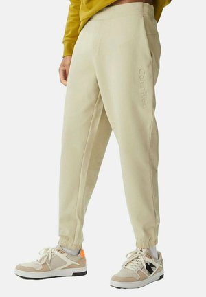 Pantalones de chándal beige con puños elásticos que cuentan con un bolsillo lateral y una marca en relieve de "Calvin Klein". Combinados con zapatillas beige y blancas.
