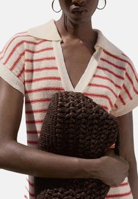 Personne portant un haut tricoté rayé crème et rouge tenant un grand sac tissé marron foncé, avec des boucles d'oreilles en forme d'anneau visibles et un fond neutre.