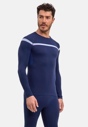Mand med krøllet hår og skæg iført tætsiddende navyblå langærmet sportstop og matchende leggings, kigger let mod venstre.