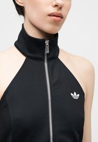 Personne portant un haut noir sans manches avec un col montant et une fermeture éclair argentée à l'avant, arborant un logo Adidas blanc sur la poitrine.