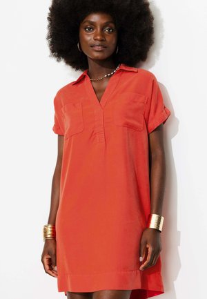 Femme avec coiffure afro portant une robe orange à manches courtes avec des poches poitrine et des bracelets dorés aux deux poignets sur fond blanc.