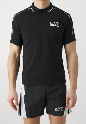 Muž nosící černé polo tričko EA7 a černé a bílé šortky EA7 stojí s uvolněnýma rukama podél těla na jednobarevném pozadí.