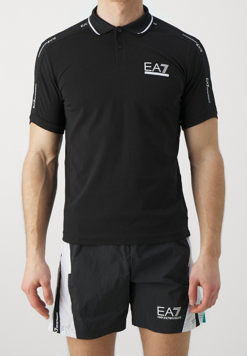 Homme portant un polo noir EA7 et un short EA7 noir et blanc, debout les bras détendus le long du corps sur un fond uni.