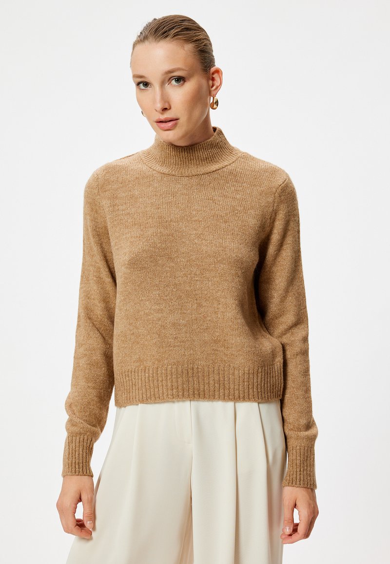Beige turtleneck neule, joka on valmistettu pehmeästä, tekstuurilla varustetusta kankaasta. Neulassa on ribbausmateriaalia hihansuissa ja helmassa, ja se on hieman lyhyt.