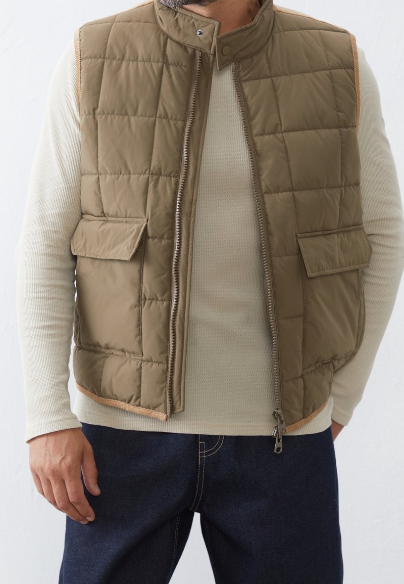 Uomo che indossa un gilet imbottito oliva su una maglietta a maniche lunghe beige in maglia waffle e jeans blu scuro, in piedi davanti a uno sfondo semplice.