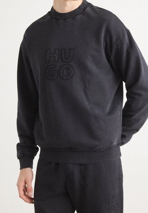 Zwarte sweatshirt met ribgebreide kraag en manchetten, voorzien van een reliëf "HU GO" logo op de borst, gemaakt van een zachte textuurstof.