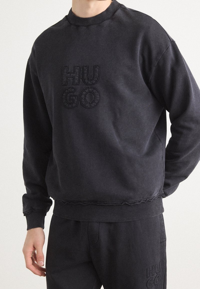 Sudadera negra con cuello y puños acanalados, con el logo en relieve "HU GO" en el pecho, fabricada con un tejido suave y texturizado.