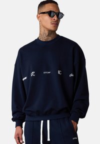 Sweatshirt bleu marine en mélange de coton, arborant divers logos blancs sur le devant. Coupe ample avec un col et des poignets côtelés.