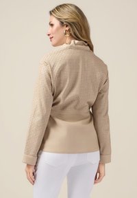 Beżowa perforowana kurtka z gładką dolną częścią i kołnierzem, długimi rękawami oraz dopasowanym krojem, w zestawieniu z białymi spodniami.