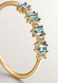 Anillo de oro que presenta gemas rectangulares alternas en tonos aqua y claras, engastadas en garras a lo largo de una banda lisa y pulida.