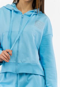Felpa leggera azzurra con zip, realizzata in tessuto morbido, dotata di fondo e polsini a coste, caratterizzata da una tasca frontale e un logo in strass sul petto.