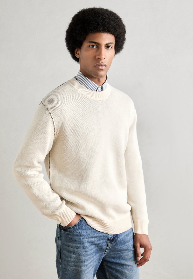 Suéter de punto color crema con cuello redondo, usado sobre una camisa de rayas azul y blanca. Combinado con jeans de mezclilla azul claro.