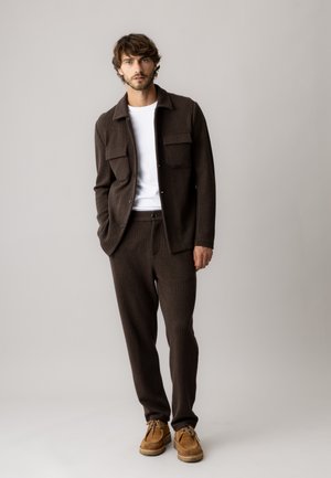 Man wearing a brown knit jacket and pants with a white shirt and brown suede shoes, standing against a plain light background.