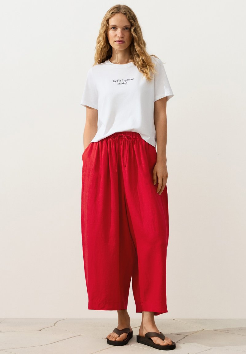 Femme aux cheveux blonds ondulés portant un t-shirt blanc avec du texte, un pantalon rouge ample et des sandales noires à plateforme, debout devant un fond uni.