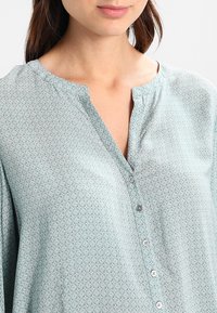 Chemise bleu clair à boutonnage, fabriquée en tissu doux, avec un motif floral, un col mao et de petits boutons sur le devant.