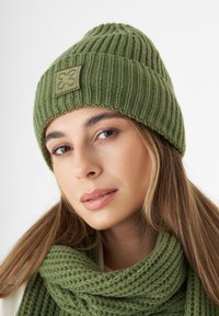 Olijfgroen geribde breibeanie met een gestructureerd patroon en een rechthoekig label. Gecombineerd met een bijpassende gebreide sjaal.