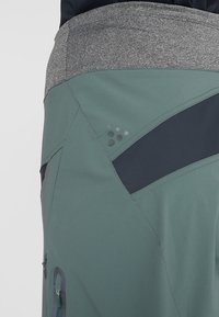 Gros plan sur un pantalon d'extérieur vert et gris avec une poche zippée et des surpiqûres renforcées sur le côté et la ceinture.