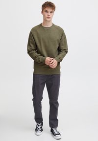 Sweatshirt verde măsliniu cu guler rotund, manșete și tiv cu zăbrele. Purtați cu pantaloni gri închis și adidași negri înalți. Design simplu.