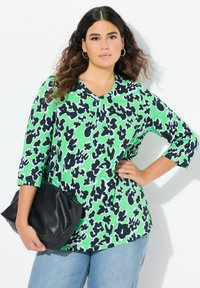 Zielona bluzka z czarnym kwiatowym wzorem, dekolt w serek i rękawy o długości 3/4, połączona z czarną kopertówką i jasnoniebieskimi dżinsami. Gładka tekstura materiału.
