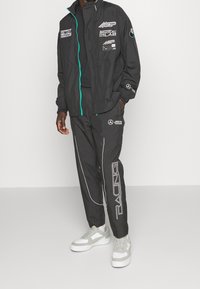 Svart vindjacka med turkos dragkedja och flera logotyper, matchad med svarta byxor med vita detaljer och texten "RACING". Grå sneakers.