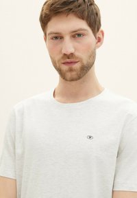 Lysegrå bomulds-t-shirt med rund hals, korte ærmer og et lille sort logo på venstre bryst. Ensartet tekstur.
