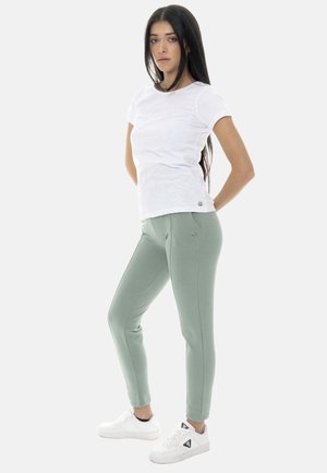 Jeune femme aux longs cheveux foncés, portant un t-shirt blanc, un pantalon vert clair ajusté et des baskets blanches, debout les mains dans le dos.