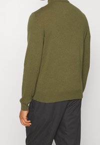 Mies, der einen olivgrünen Langarmpullover und lockere schwarze Hosen trägt, steht mit dem Rücken zu einem schlichten weißen Hintergrund.