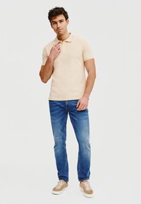 Beżowa koszulka polo, slim fit, wykonana z bawełny. Noszona z niebieskimi dżinsami, jasny odcień. Obuwie: brązowe sneakersy, sznurowane.