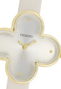 Montre à bracelet en cuir blanc avec un boîtier plaqué or en forme de trèfle à quatre feuilles, un cadran argenté, des aiguilles dorées et le nom de la marque "OOZOO" affiché.