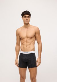 Fekete boxer alsónadrág geometrikus mintázattal, elasztikus Calvin Klein derékkal, és testhez simuló szabásban, férfi modellen bemutatva.