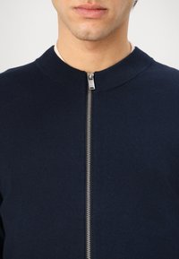 Námornícky zipsový sveter s ribbed golierom, hladkou textúrou a detailom strieborného kovového zipsu. Detailný pohľad zameraný na oblasť hrudníka.