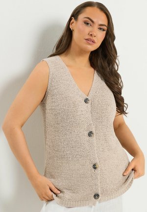 Donna con lunghi capelli castani che indossa un gilet beige senza maniche lavorato a maglia con bottoni, in piedi contro una parete chiara, con le mani nelle tasche.