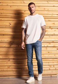 Hombre con tatuajes en ambos brazos, vistiendo una camiseta blanca, jeans azules y zapatillas blancas, se apoya contra una pared de tablones de madera.