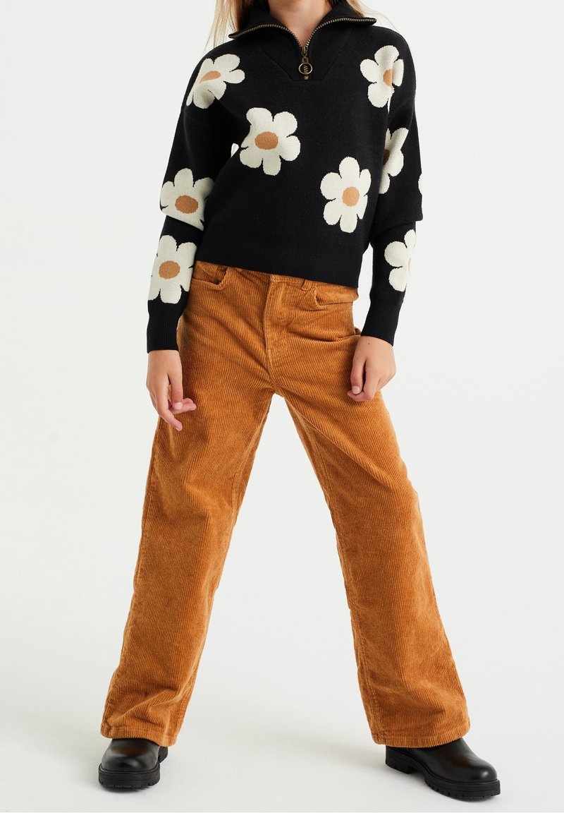 Enfant portant un pull noir avec des motifs de fleurs blanches et beiges, un pantalon en velours côtelé marron et des bottes noires, debout jambes écartées.