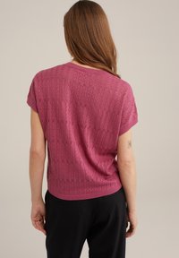 Růžový pletený top s krátkými rukávy, zdobený vzorem s průstřihy a žebrovaným lemem. Následuje uvolněnou siluetu nošený s černými kalhotami.