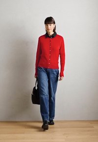 Red knitted cardigan with buttons, worn over a black collared shirt, paired with blue jeans and black shoes. Model holds a black handbag.