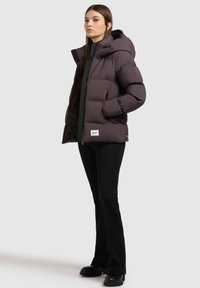 Chaqueta acolchada morada oscura con capucha, que cuenta con un cierre frontal, dos bolsillos laterales y un parche de logo en el dobladillo. Llevada sobre pantalones negros.