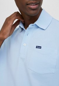 Jasnoniebieska koszulka polo z teksturowanej tkaniny, z kołnierzykiem, dwiema guzikami oraz kieszonką na piersi z ciemnoniebieską metką.