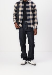 Man in een marineblauw en beige geblokte shirt over een zwart T-shirt, donkere jeans, witte sneakers, en met een zwarte schoudertas tegen een witte achtergrond.