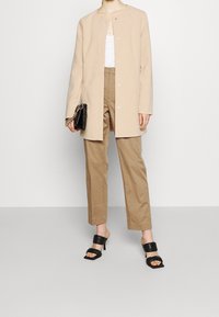 Cappotto beige a maniche lunghe con bottoni a pressione, abbinato a pantaloni beige e mule nere con tacco. Gli accessori includono una piccola borsa nera.