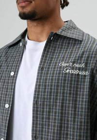 Knopfleiste mit Hemd in dunkelgrünem und marinekarriertem Muster mit weißer Stickerei mit der Aufschrift "Can't rush Greatness", mit klassischem Kragen und Knöpfen.