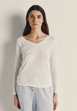 Jauna sieviete ar gariem tumšiem matiem baltā garroku v-veida kakla kreklā un gaiši pelēkās sporta biksēs, stāvot pret vienkāršu fonu.