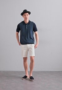 Polo majica s kratkimi rokavi z navy vzorcem spredaj, temnima rokavoma, v kombinaciji z belimi kratkimi hlačami in črnimi espadrili. Model nosi črno klobuk.