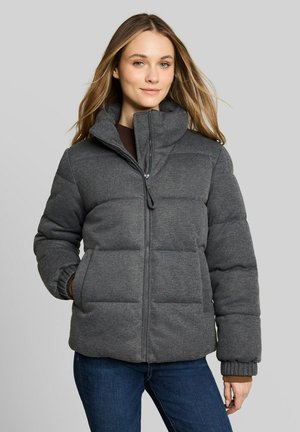 Frau trägt eine dunkle, grau gepolsterte Jacke mit Reißverschluss, hohem Kragen und elastischen Bündchen, kombiniert mit blauen Jeans, steht vor einem einfarbigen Hintergrund.