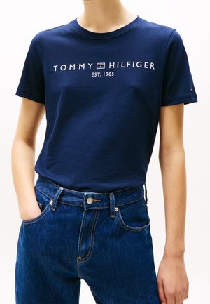 Osoba nosząca granatowy t-shirt Tommy Hilfiger z logo oraz ciemnoniebieskie jeansy, jedną ręką w kieszeni, na białym tle.