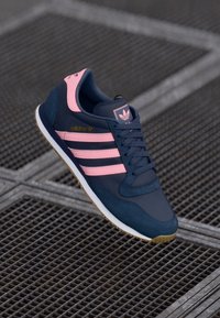 Shoe atlético azul marinho com detalhes em rosa, três riscas e logótipo dourado. Apresenta um cabedal em camurça texturizada e sola de borracha.