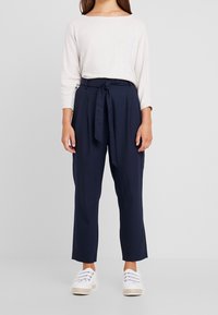 Pantalon taille haute bleu marine avec un nouage frontal, associé à un haut à manches longues beige clair et des baskets blanches. Tissu à texture lisse, coupe décontractée.