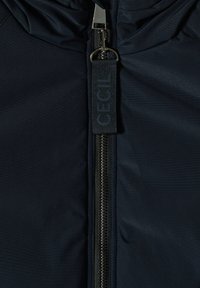 Giacca blu navy in tessuto liscio e opaco; presenta una zip frontale e un'etichetta con logo vicino al colletto per identificazione.