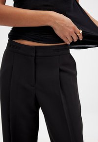 Juodos pritaikytos kelnės su lygiu paviršiumi, papuoštos priekinėmis raukšlėmis ir subtiliais šoninio siuvimo akcentais, suteikiančiais struktūrizuotą išvaizdą.